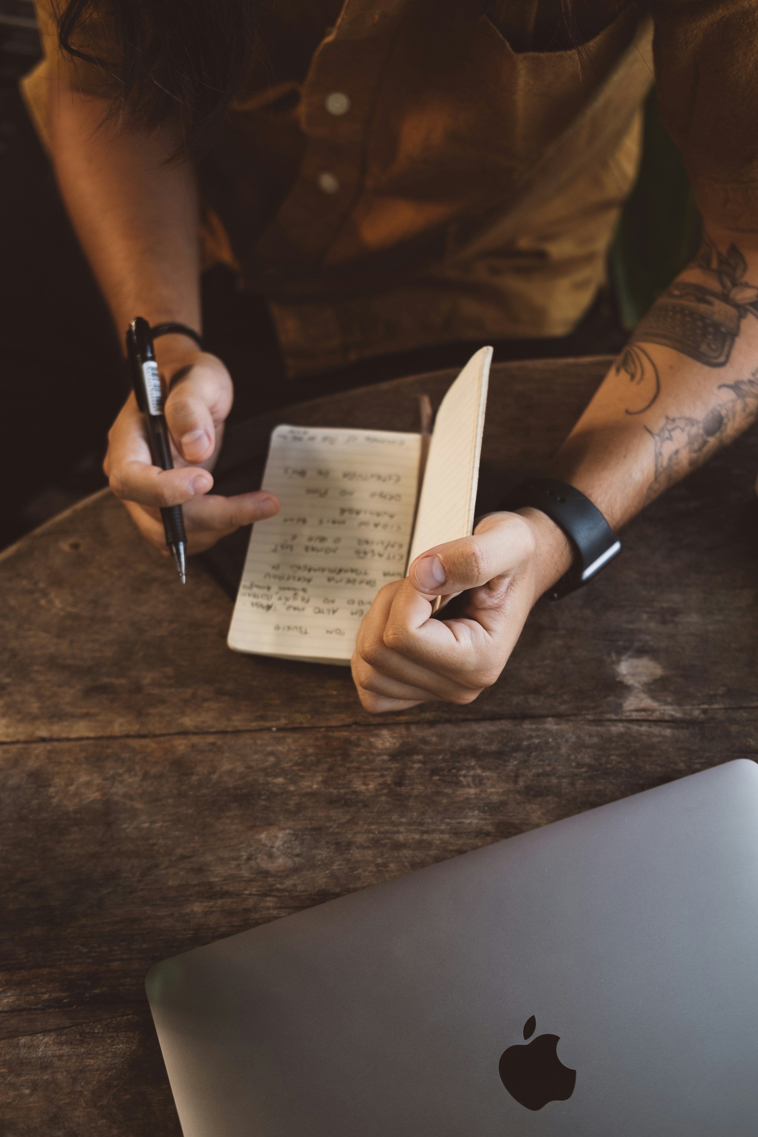 Person writing on a notepad with a pen, next to an Apple laptop on a wooden surface.